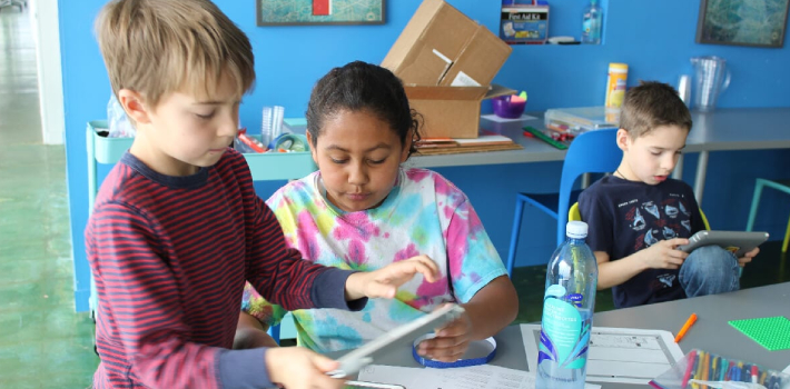 Two students are working together on a project in school. They are looking into a tablet. Some papers are kept on the desk. Another boy is sitting nearby watching his gadget.